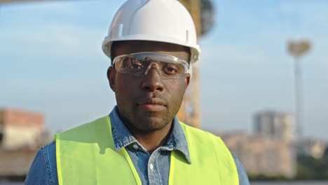 Nahaufnahme-Des-Jungen,-Gutaussehenden-Afroamerikanischen-Bauunternehmers-Mit-Schutzhelm-Und-Schutzbrille,-Der-In-Die-Kamera-Blickt-Und-Lächelt.-Outdoor-Auf-Der-Baustelle.